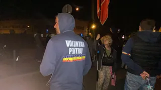 Un bărbat cu o hanorac albastru pe care scrie un text colorat stă cu spatele, în timp ce alți oameni se află în jur pe timp de noapte.