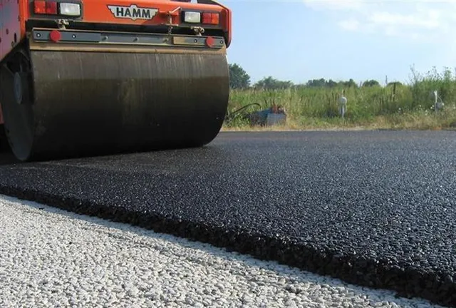 Un aparat de compactare cu cilindru metalic parcurge o suprafață de asfalt proaspăt întinsă pe un drum de pietriș.
