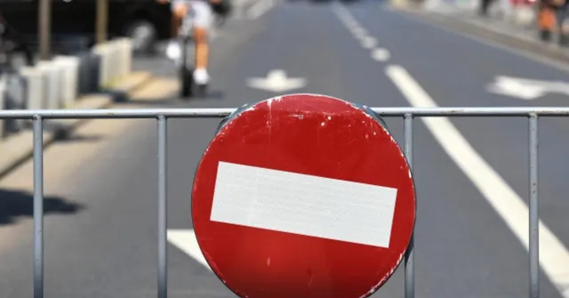 Un semn de circulație circular, roșu cu o bandă albă orizontală, este montat pe un gard metalic gri în fața unei străzi asfaltate.