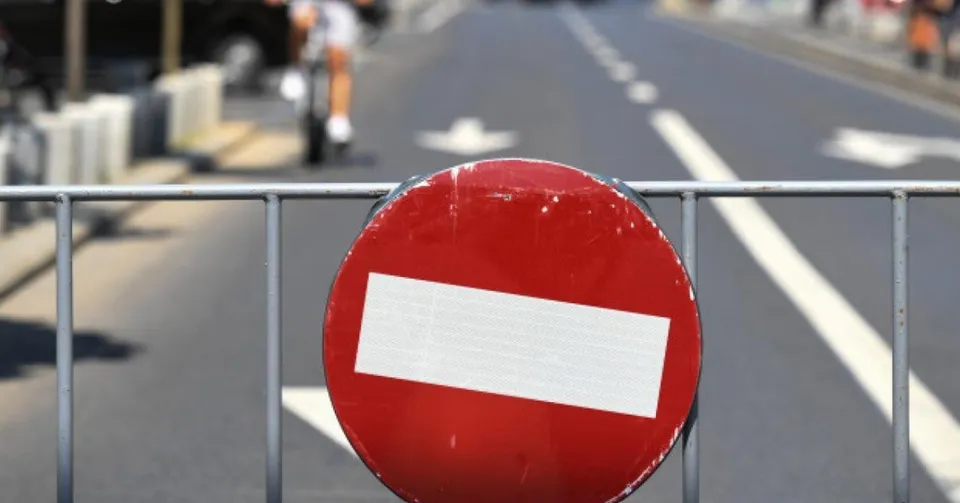 Un semn de circulație circular, roșu cu o bandă albă orizontală, este montat pe un gard metalic gri în fața unei străzi asfaltate.