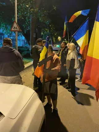 Un grup de persoane stă pe un drum lângă mai mulți oameni care țin steaguri colorate sub lumina unui stâlp de iluminat nocturn.