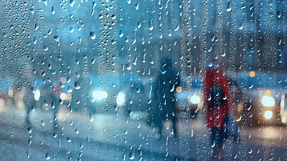 Picături de apă se scurg pe o suprafață transparentă, prin care se observă siluete de oameni și lumini de mașini pe un fundal urban întunecat.