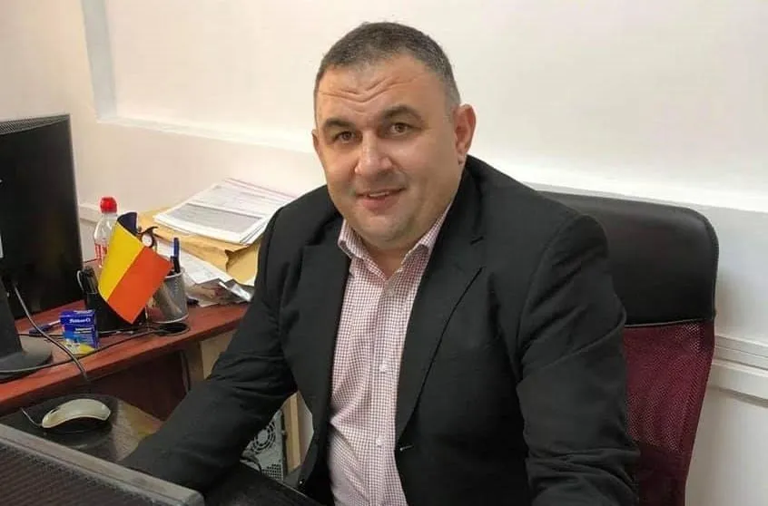 Un bărbat cu sacou negru și cămașă roz cu model este așezat la un birou lângă un monitor și un steag.