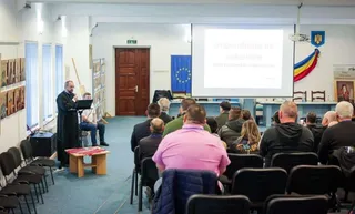 O persoană în costum negru vorbește la un microfon în fața unei mulțimi de oameni care se uită la un ecran de proiecție într-o sală de curs.