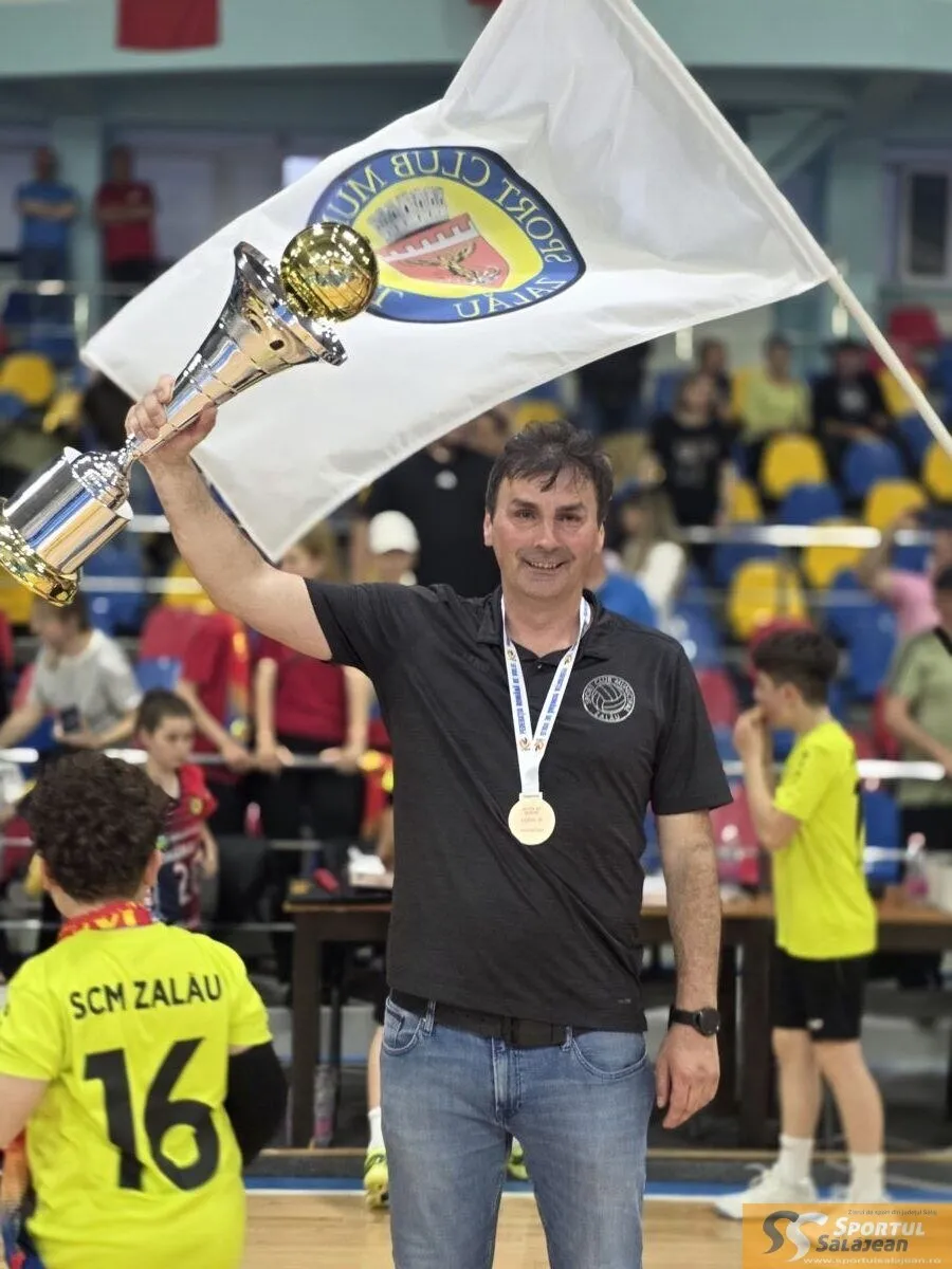 Un bărbat cu medalie aurie și tricou negru ridică un trofeu și un steag alb într-o sală de sport.