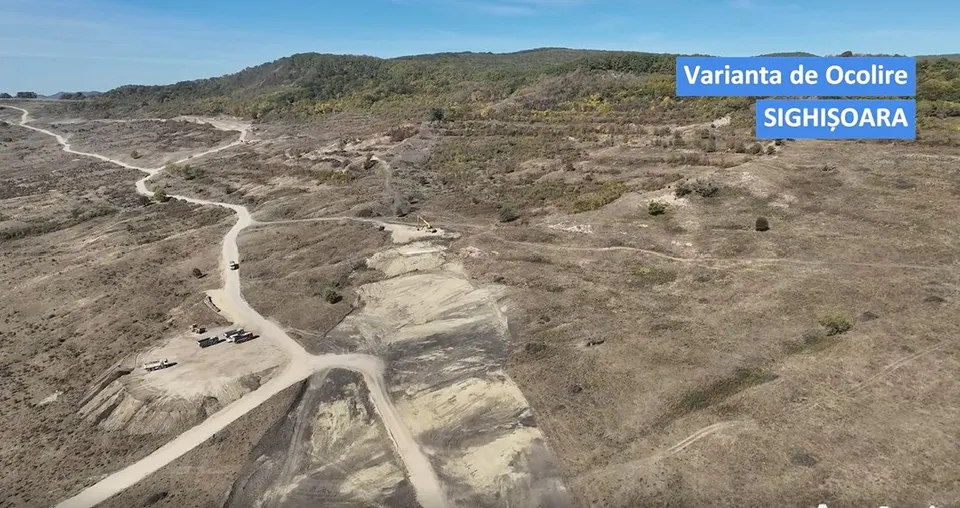 O vedere aeriană prezintă un teren accidentat cu drumuri de pământ și zone de excavare sub un cer senin.