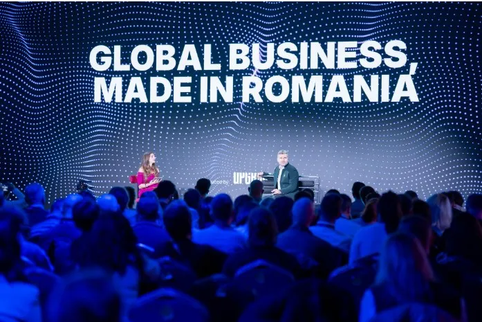 O mulțime de oameni stă în fața unei scene unde o femeie și un bărbat se află sub un ecran mare cu text alb pe fundal albastru.