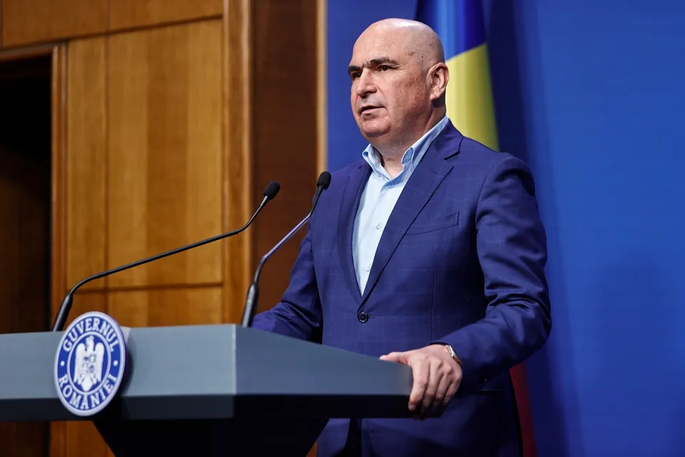 Un bărbat cu pielea deschisă, purtând sacou albastru și cămașă deschisă la guler, stă în spatele unui podium gri cu un emblema circulară.