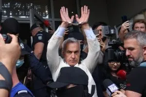 Un bărbat cu părul cărunt și haine deschise la culoare poartă cătușe la mâinile ridicate în timp ce este înconjurat de microfoane și camere.