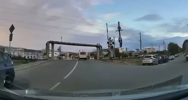 Privire de pe bordul unei mașini spre o trecere la nivel cu bariere ridicate și un vehicul alb circulând pe drumul asfaltat.