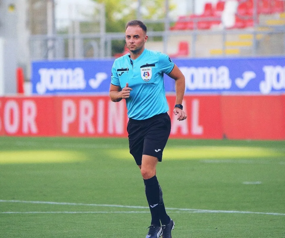 Un bărbat cu tricou albastru și pantaloni scurți negri aleargă pe un teren de fotbal verde în fața unor panouri publicitare colorate.