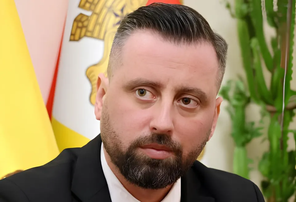 Un bărbat cu barbă și păr scurt stă în fața unui steag și al unei plante verzi.