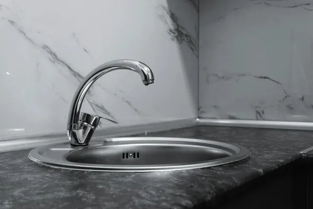 Un robinet metalic este montat pe o chiuvetă de inox într-un colț de bucătărie cu blat închis și perete marmorat.
