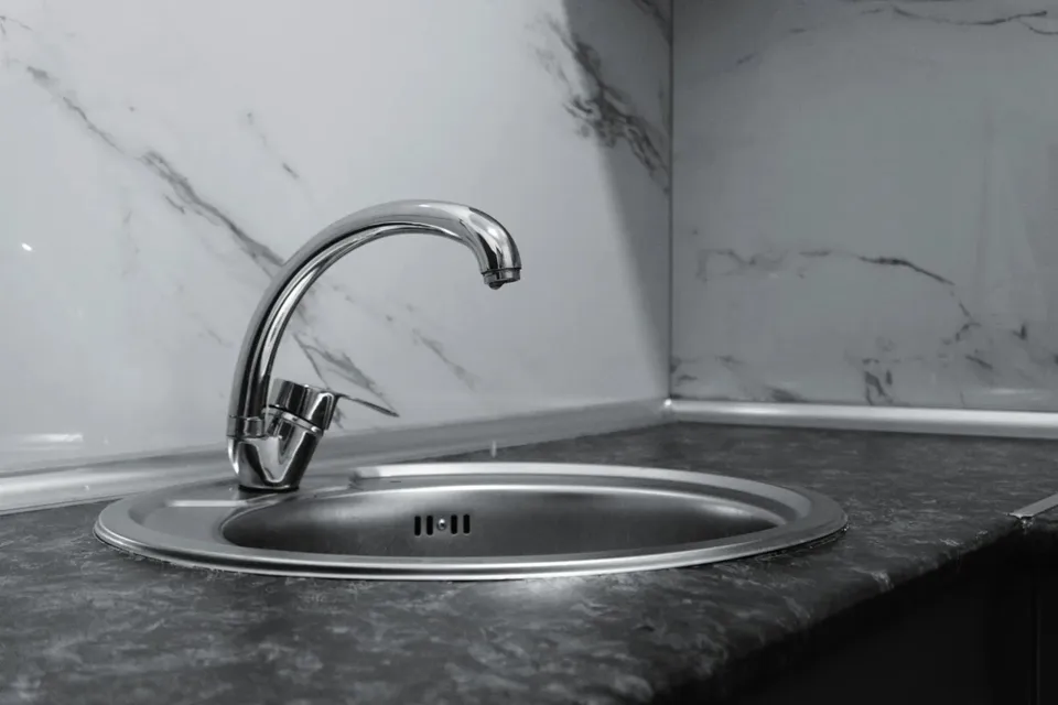 Un robinet metalic este montat pe o chiuvetă de inox într-un colț de bucătărie cu blat închis și perete marmorat.