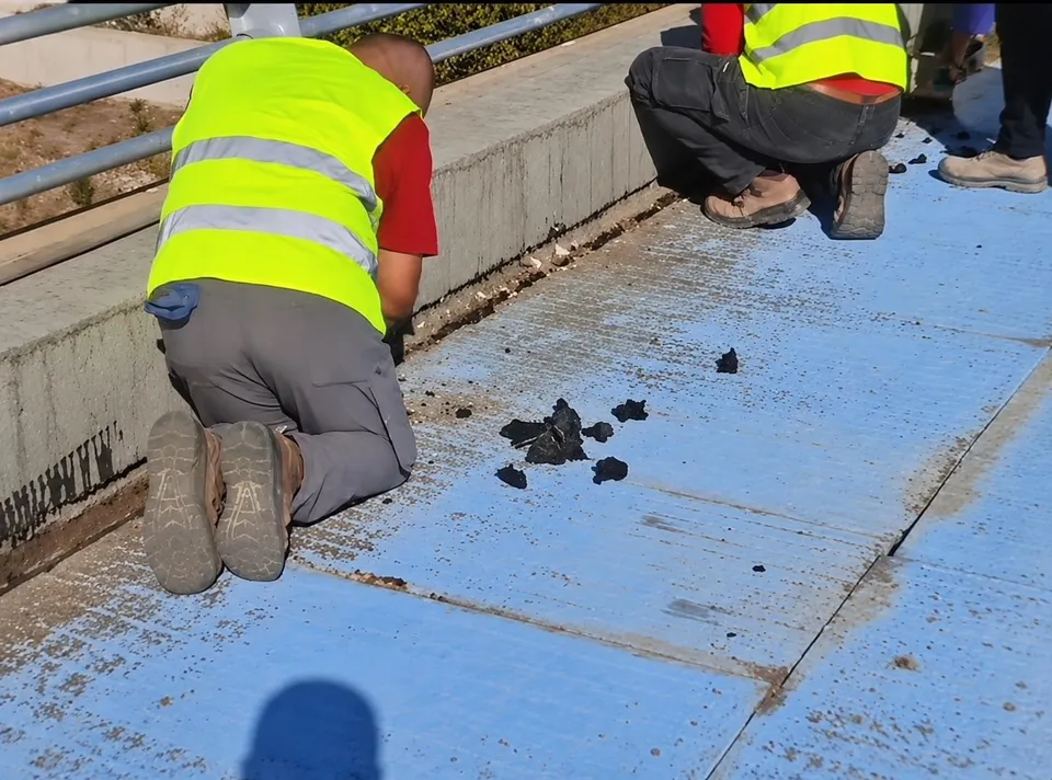 Două persoane purtând veste fluorescente galben-verzi stau în genunchi pe o suprafață albastră, inspectând fragmente negre de material împrăștiate pe sol.