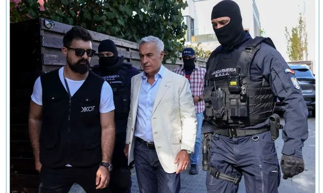 Un bărbat cu sacou deschis merge pe stradă înconjurat de un bărbat cu vestă neagră și de doi agenți în uniformă de poliție și măști negre.