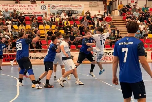 Câțiva jucători de handbal în echipamente albastre și albe se află pe un teren de sport în fața unor spectatori așezați în tribune.