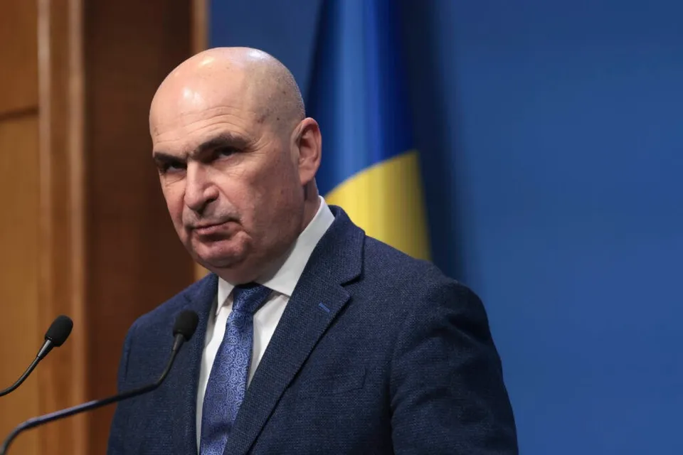 Un bărbat cu capul chel, purtând sacou albastru, cămașă albă și cravată albastră, stă în fața a două microfoane.