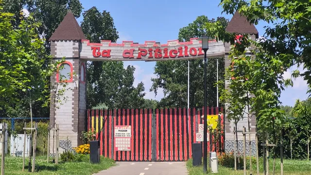 O poartă de intrare cu turnuri și un panou cu text roșu se află în fața unui gard cu bare verticale roșii și negre.