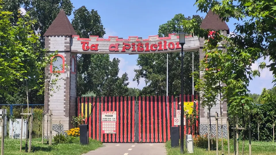 O poartă de intrare cu turnuri și un panou cu text roșu se află în fața unui gard cu bare verticale roșii și negre.