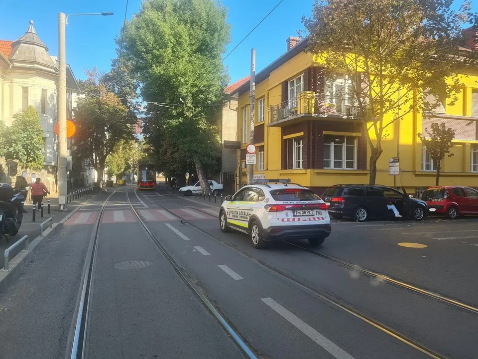 O mașină albă cu inscripții roșii și albastre circulă pe un drum cu șine de tramvai, lângă clădiri și copaci sub un cer senin.