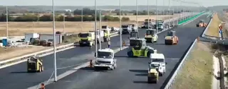 Mai multe utilaje de construcții și camioane circulă pe o porțiune de drum asfaltat aflată în lucru.