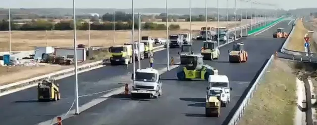 Mai multe utilaje de construcții și camioane circulă pe o porțiune de drum asfaltat nou, lângă un teren deschis sub un cer senin.