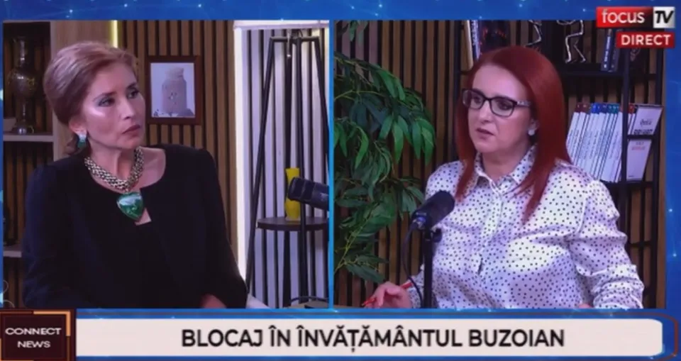 Două femei stau în cadru, una purtând o bluză neagră cu un colier mare, iar cealaltă o cămașă albă cu puncte negre și ochelari.