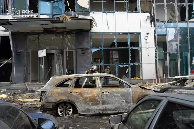 O mașină arsă stă în prim-plan în fața unei clădiri cu geamuri sparte și fațadă deteriorată.