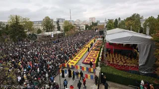 O mulțime mare de oameni stă adunată într-un spațiu deschis lângă o scenă acoperită și un drum decorat cu flori colorate.