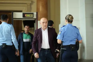 Un bărbat cu sacou purpurii și cămașă albă stă între doi polițiști în uniformă albastră într-un spațiu cu un detector de metale în fundal.