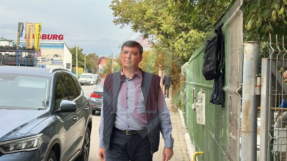 Un bărbat cu vestă închisă și cămașă în dungi pășește pe un trotuar lângă mașini și un gard verde.