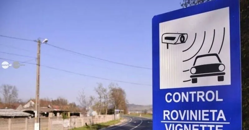 Un panou albastru de tip control electronic este vizibil lângă o cale rutieră, cu o cameră și un autovehicul desenate pe el.