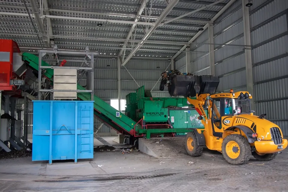 Un tractor galben cu braț hidraulic este poziționat în fața unor utilaje industriale verzi și a unui container albastru într-un spațiu mare acoperit.