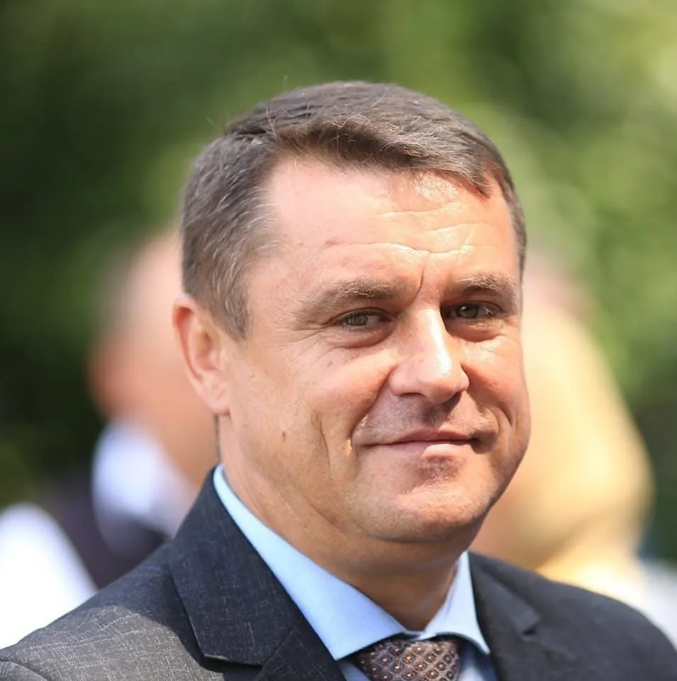 Un bărbat cu părul scurt și cămașă albastră poartă un sacou închis la culoare și o cravată în timp ce privește spre camera foto.