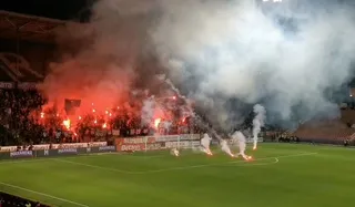 Fumul dens de culoare albă și roșie acoperă o parte din terenul de fotbal și tribunele unui stadion noaptea.