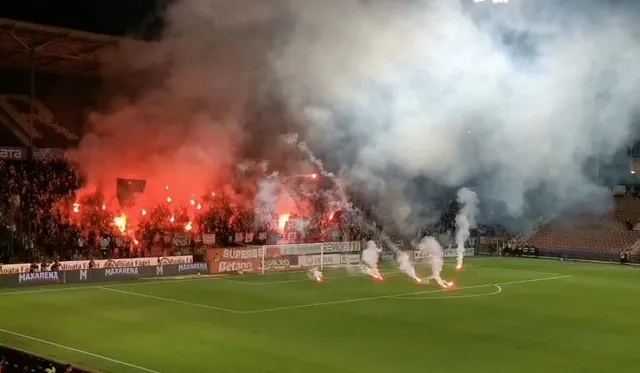 Fumul dens de culoare albă și roșie acoperă o parte din terenul de fotbal și tribunele unui stadion noaptea.