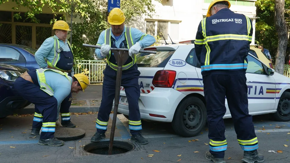 Patru muncitori cu căști galbene și veste reflectorizante lucrează lângă un gura de canal deschisă pe un drum lângă mașini parcate.