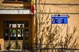 O clădire cu o intrare glaspă și un panou albastru pe peretele exterior, sub care se proiectează umbre de ramuri de copaci.