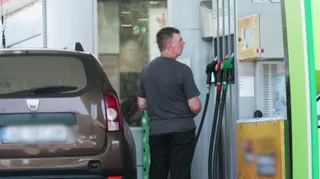 Un bărbat stă lângă o pompă de benzină lângă partea din spate a unei mașini maronii.