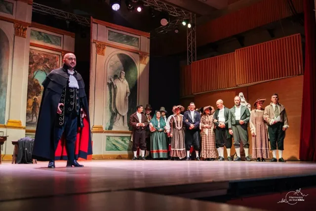 Un grup de actori în costume de epocă stă în linie pe o scenă de teatru în fața unui decor pictat.