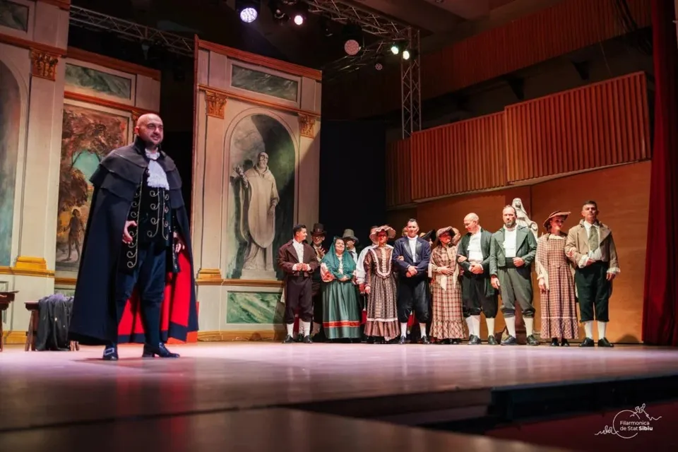 Un grup de actori în costume de epocă stă în linie pe o scenă de teatru în fața unui decor pictat.