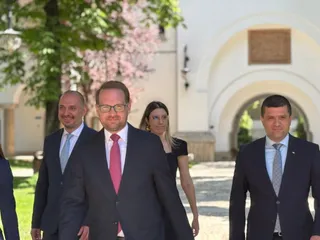 Patru persoane îmbrăcate în costume și haine office merg pe un drum pavat lângă o clădire albă cu arcade și copaci verzi.