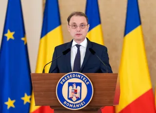 Un bărbat cu ochelari și cravată stă în spatele unui podium de lemn pe care este afișat un emblema circulară, în fața unor steaguri colorate.