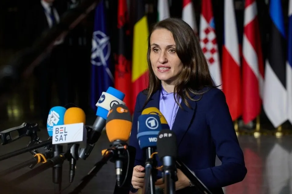 O femeie cu părul lung și sacou albastru stă în fața mai multor microfoane și a unor steaguri colorate.