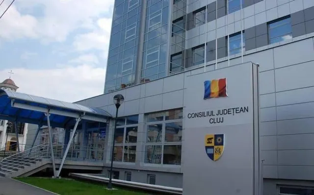 O clădire modernă cu fațadă de sticlă și panouri gri prezintă un panou cu textul Consiliul Județean Cluj și un blazon colorat.