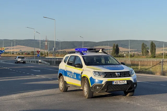 Un vehicul de poliție alb cu marcaje galbenii și albastre este parcat pe o porțiune de drum asfaltat sub un cer senin.