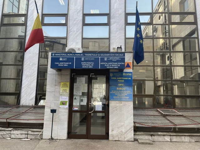 Clădire cu intrare centrală, ferestre mari și două steaguri română și UE așezate pe stâlpi laterali.