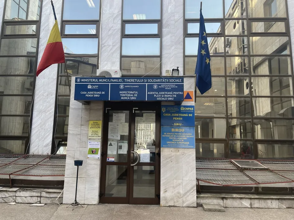 Clădire cu intrare centrală, ferestre mari și două steaguri română și UE așezate pe stâlpi laterali.