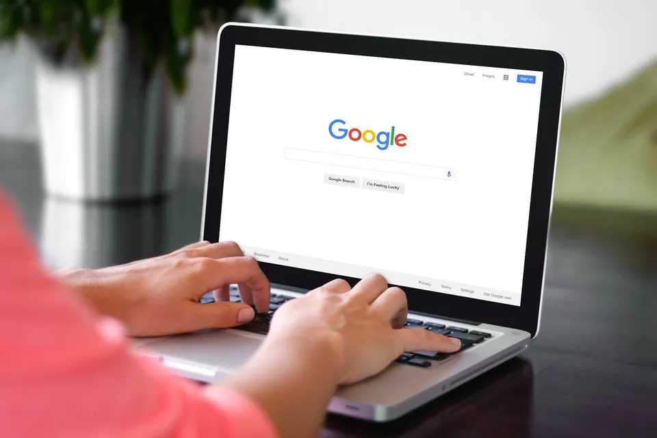 O persoană în magiliu roz tastează pe un laptop aflat pe o masă întunecată, afișând pagina principală Google pe ecranul dispozitivului.